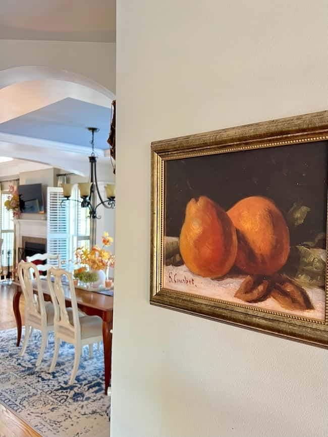 A decorative painting of two pears in a golden frame hangs on a cream-colored wall. In the background, a dining area with a wooden table, white chairs, and chandelier comes into view, exuding warm autumn refresh vibes. The room is well-lit with natural light and offers simple decor ideas for a cozy fall home tour.