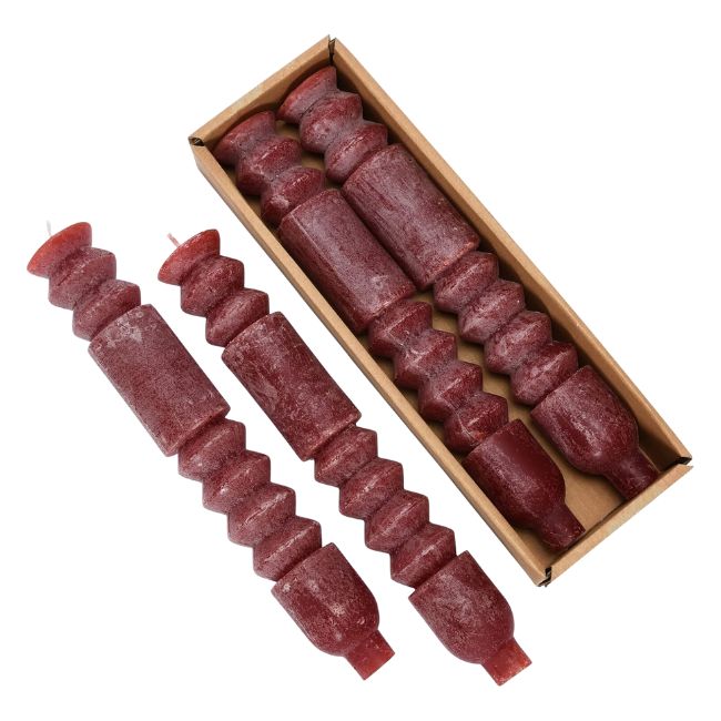 A set of six long, intricately-shaped reddish brown candles, four of which are in a rectangular cardboard box. Two candles are placed outside the box. Each candle has a unique pattern with various ridges and segments along its length.