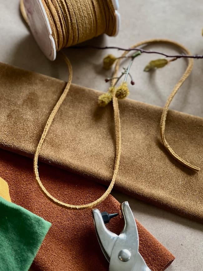 Craft supplies including spools of brown and green fabric, a piece of yellow suede cord, plant sprigs, and a metal hole punch tool are arranged on a table.