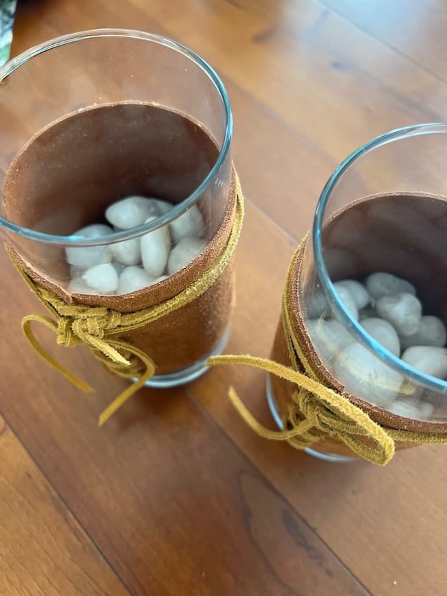 Two glass vases wrapped with brown fabric and tied with a yellow ribbon, each filled with white stones, are placed on a wooden surface. The vases have a rustic decorative look.