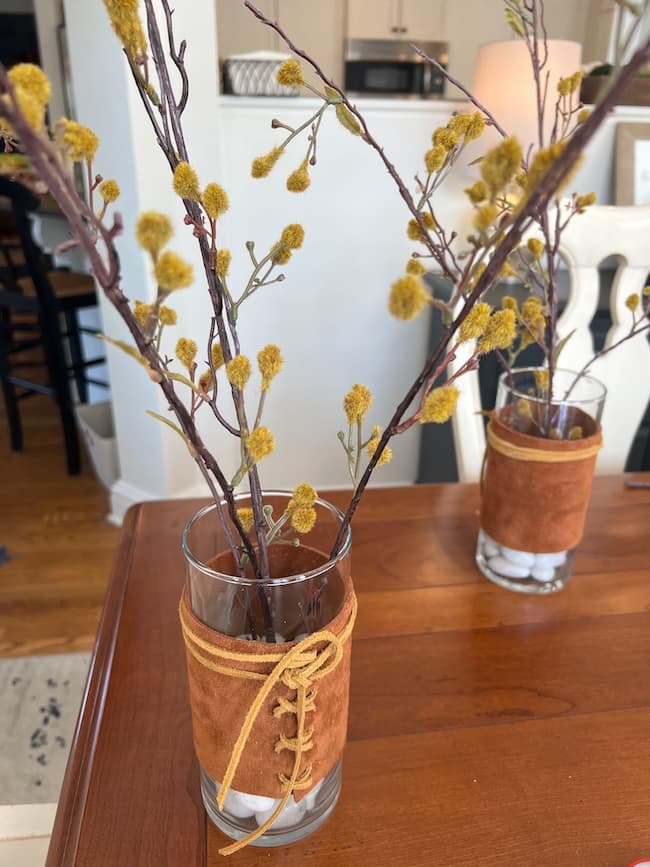 Glass vases with brown suede wraps and lace-up details stand on a wooden table. Each vase contains thin branches with small yellow blossoms. A blurred background suggests a cozy indoor setting.