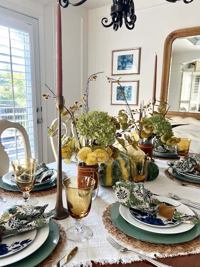 A dining table set for four with green and white dishes, floral napkins, amber glasses, and tall brown candles. The centerpiece features yellow and green squashes, greenery, and hydrangeas. The room has white walls, a mirror, and framed artwork.