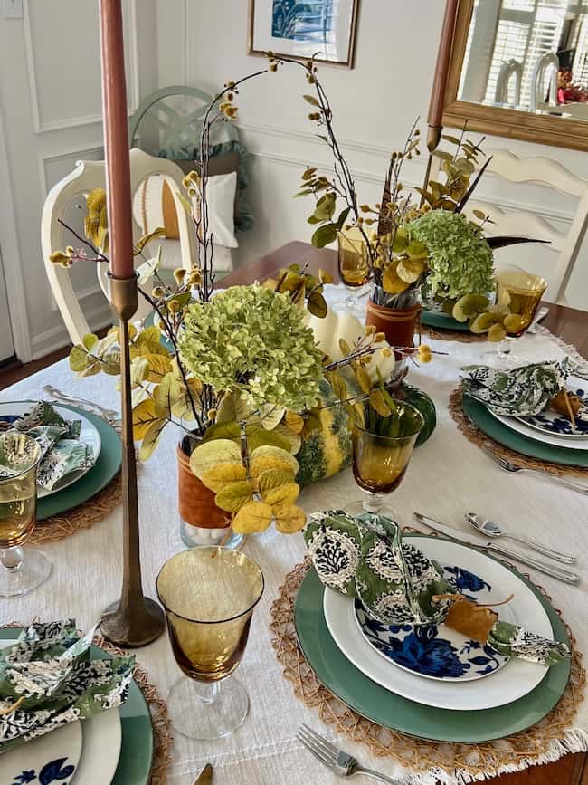 A dining table is elegantly set with green and white plates, floral napkins, and amber glassware. A white tablecloth with woven placemats complements the arrangement. Centerpieces include green and yellow flowers with tall, thin candles.