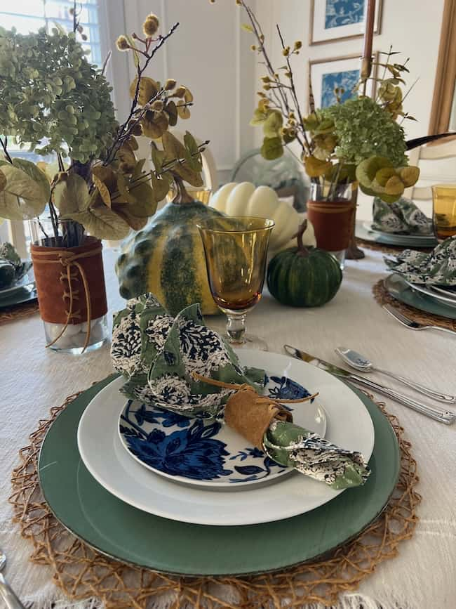 A dining table is set with a green and blue floral plate, cloth napkin, amber glass, and autumn-themed decor. Small pumpkins, dried flowers in vases, and DIY leather fall table accents add a seasonal touch to the elegant setup.