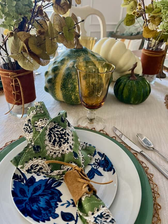 A beautifully set autumn table features a plate with a green and blue floral napkin, glassware, and a centerpiece with various squash and dried flowers. The rustic decor enhances the fall theme.