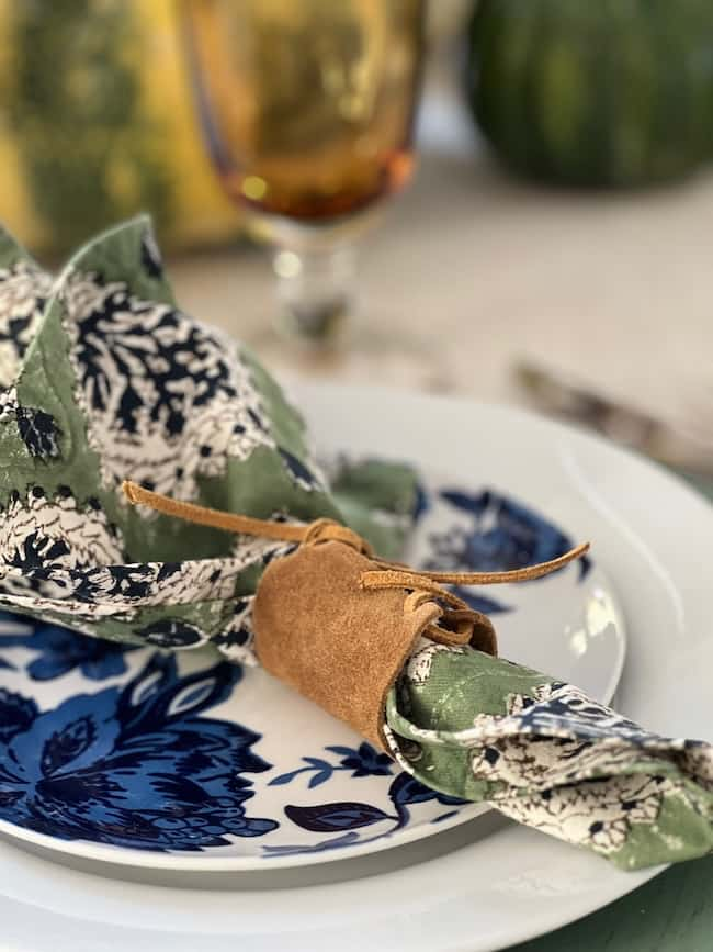 A table setting showcases DIY leather fall table decor with a blue and white floral plate paired with a green and white patterned napkin, elegantly secured by a brown leather napkin ring. In the background, a blurred glass and gourds complete the autumnal ambiance.