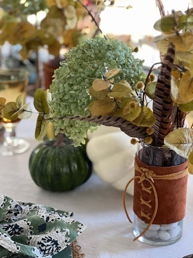 A fall-themed table centerpiece featuring a glass vase wrapped in brown fabric with lace, filled with a green hydrangea, leaves, and feathers. Nearby are decorative pumpkins and a wine glass, all set on a white tablecloth.