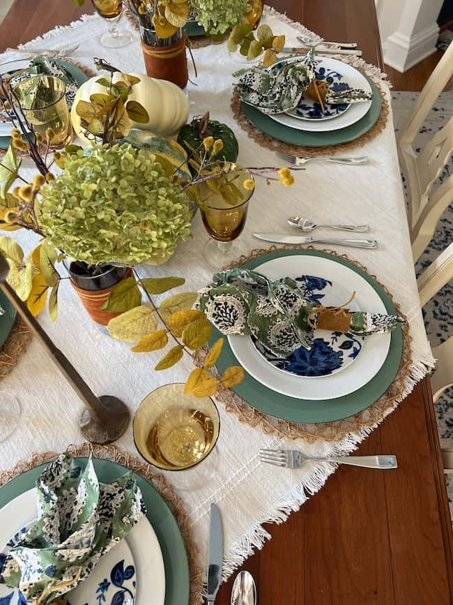 A beautifully set dining table with autumn-themed decor. It features a centerpiece of green and yellow foliage, patterned plates, fabric napkins, and golden glassware on a light tablecloth with earthy accents.