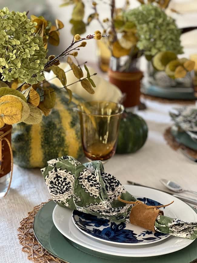 Elegant table setting with green plates, a blue and white patterned bowl, and a floral napkin held by a brown ring. The table is adorned with green and yellow floral arrangements and decorative gourds. Amber-colored glass adds warmth to the scene.