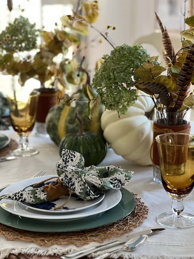 A beautifully set autumn table with plates, amber glasses, and a fabric napkin on each setting. The centerpiece includes pumpkins, gourds, hydrangeas, and foliage in earthy tones, creating a cozy and festive atmosphere.