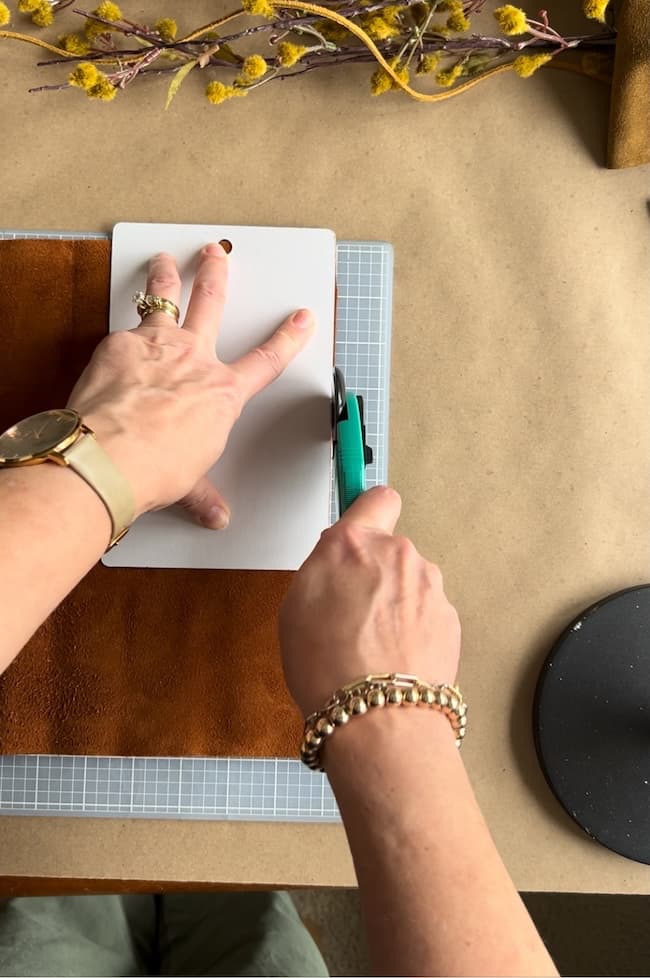 A person with a watch and bracelet uses a green utility knife to cut a piece of leather on a cutting mat. Yellow flowers and a round black object are partially visible on the table.