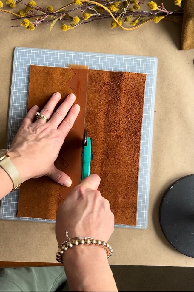 A person uses a rotary cutter to cut brown leather on a grid mat. The cutting edge has a zigzag design. There are yellow dried flowers at the top of the image. The person's hands are adorned with rings and bracelets.