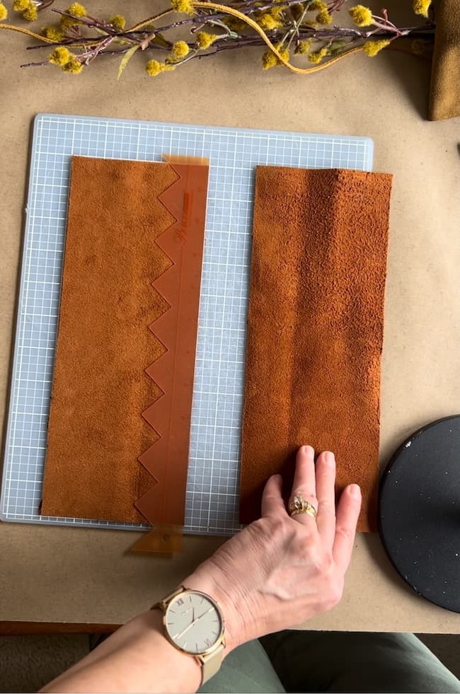 A person wearing a watch arranges two pieces of brown leather on a cutting mat. One piece features a zigzag edge. Yellow floral branches lay near the top of the mat.