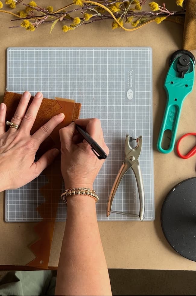 A person working on a crafting project at a table with a cutting mat, using a pen to mark on leather. Tools like a plier and plastic cutter are nearby. Yellow flowers and a small bowl are seen in the background.