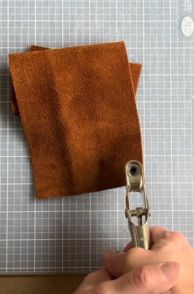 A piece of textured brown fabric rests on a gray grid-patterned cutting mat. A hand holds a metal hole punch tool above the fabric, suggesting preparation for crafting or sewing.