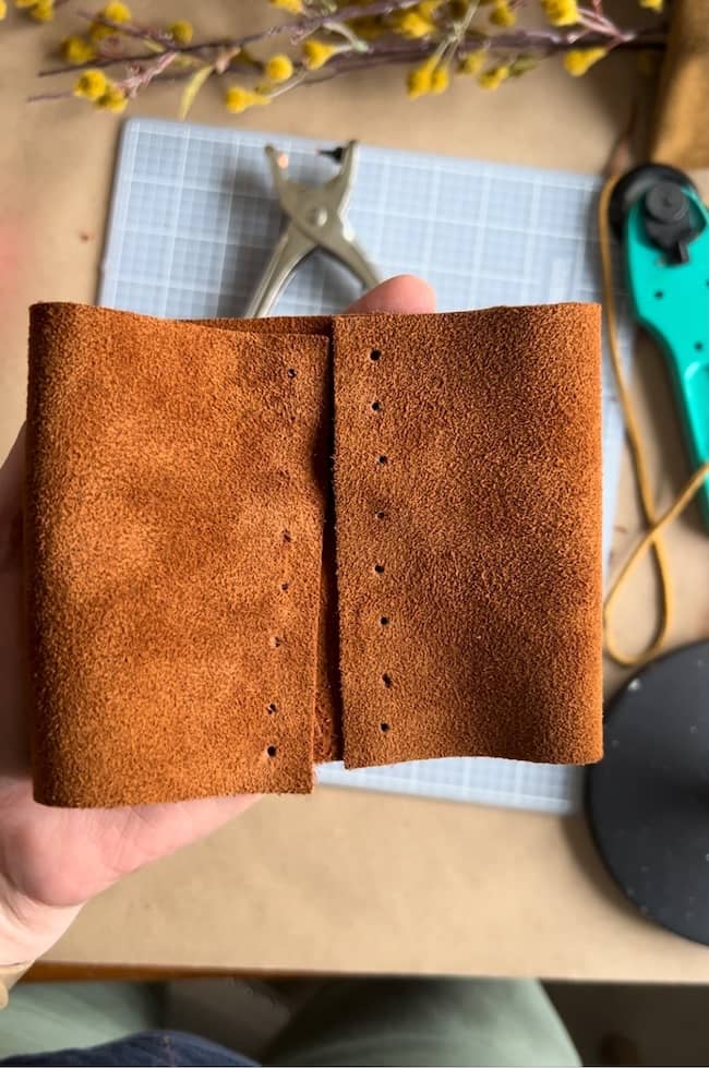 A person holds a piece of brown leather with evenly spaced holes punched along the edges. A green rotary cutter and a hole punch tool are on a grid cutting mat in the background.