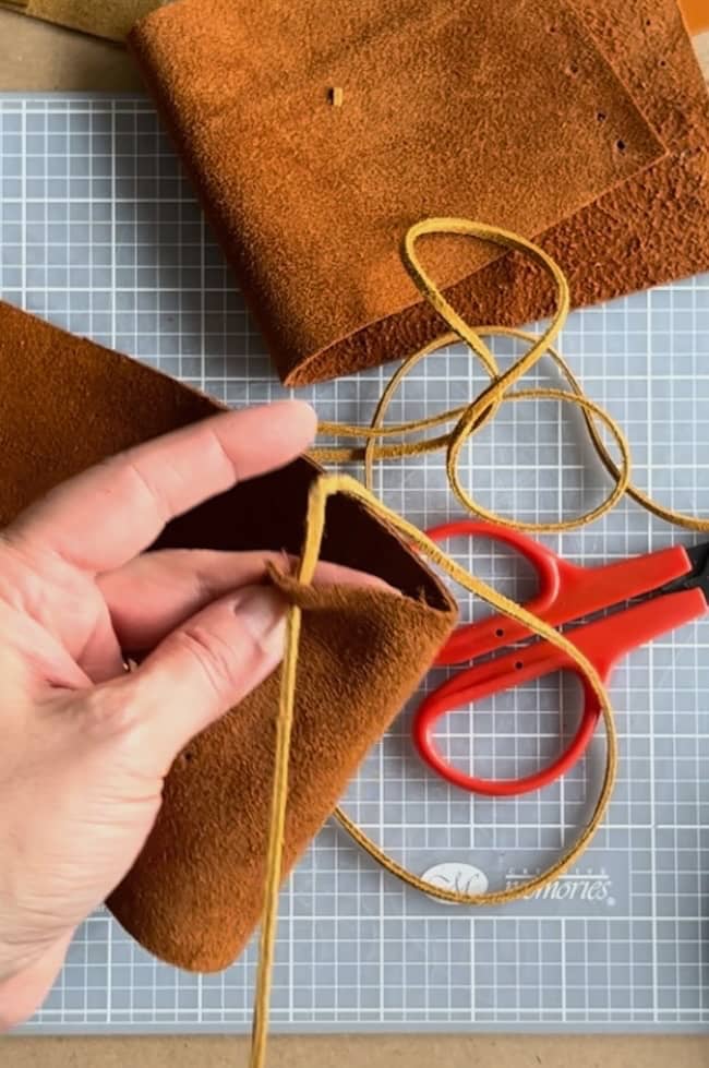 A person holding brown leather fabric with a tan cord being threaded through it. Red scissors and a rolled-up piece of leather are placed on a grid-patterned cutting mat in the background.