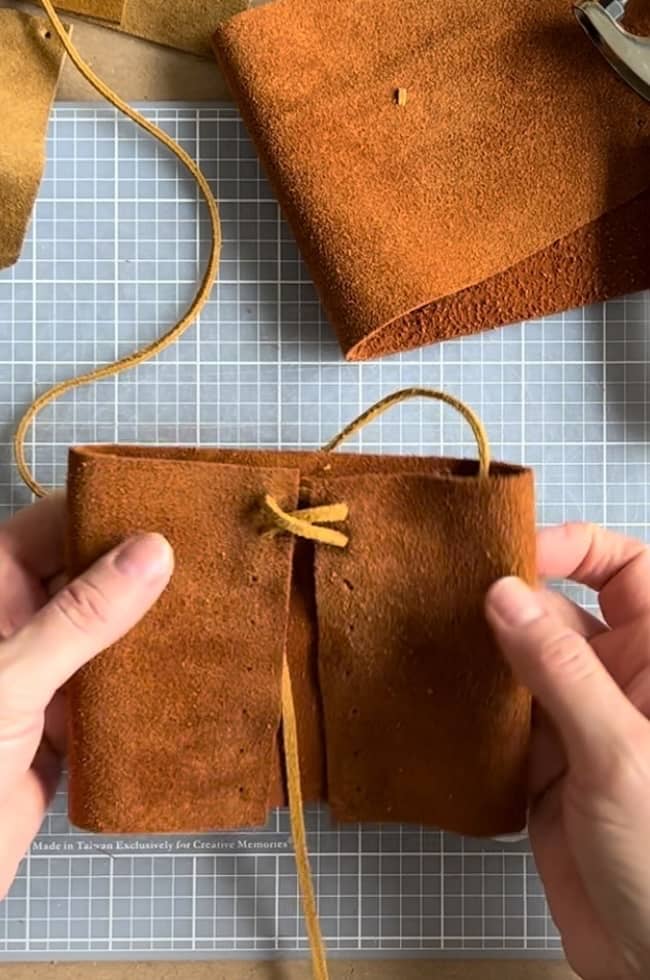 Two hands are crafting with brown leather on a gray grid cutting mat. The leather piece is being folded and laced with a thin strap. Rolls of leather are in the background.