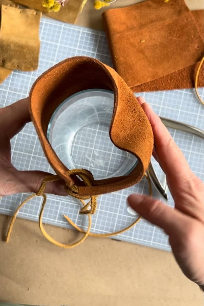 Hands wrapping a glass jar with a piece of brown leather on a crafting table. A grid mat, scissors, and pieces of leather are visible in the background.