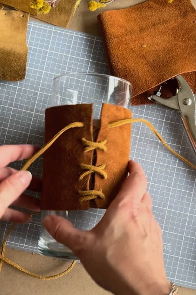 Hands are crafting a leather sleeve for a glass using brown leather cord. The sleeve features leather pieces with holes for lacing. A hole punch tool and additional leather pieces are visible in the background on a grid-patterned surface.