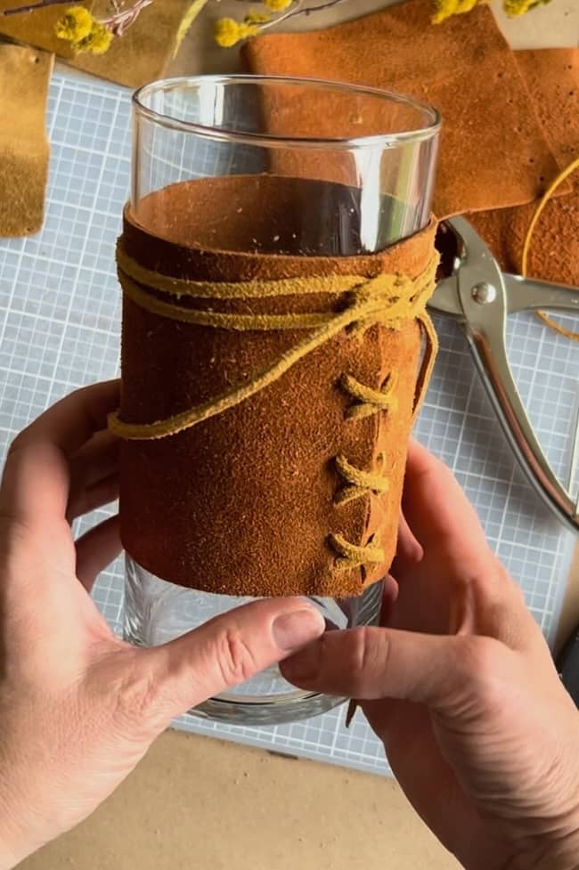 A person holds a glass wrapped in brown leather, tied with a yellow cord in a crisscross pattern. Scissors and leather pieces are scattered on a grid-patterned surface below.