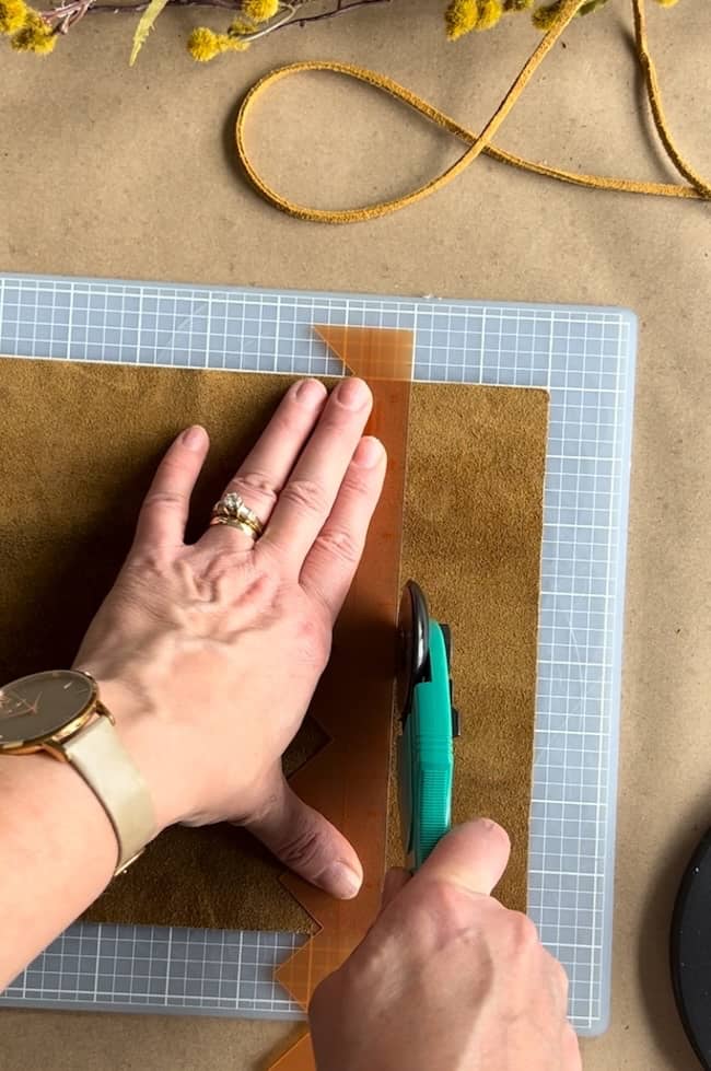 A person is using a rotary cutter and a transparent ruler to cut brown leather on a gridded cutting mat. Their left hand steadies the ruler, and they are wearing a watch and rings. A piece of fabric and ribbon are in the background.