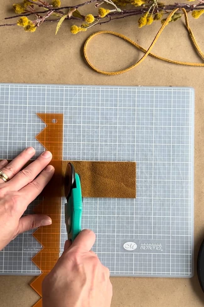 A person is using a rotary cutter and ruler on a brown piece of fabric on a grid cutting mat. Dried flowers and a piece of leather string are in the background.