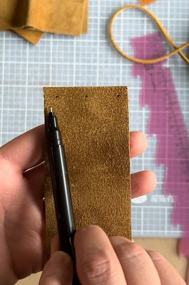 A person holds a brown leather piece with one hand and marks it with a black pen using the other hand. A transparent ruler and leather strips are visible on a cutting mat in the background.