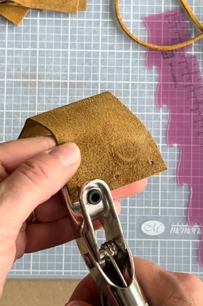 A hand holding a piece of brown leather with a metal hole punch tool, ready to create holes. A pink ruler and cutting mat are visible in the background.