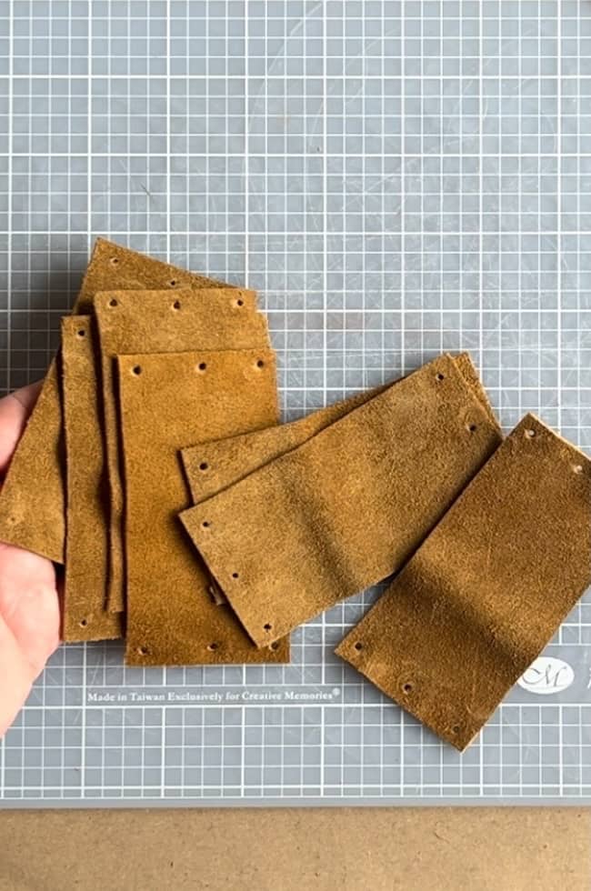 A hand holding several rectangular pieces of brown suede with small holes along the edges, placed on a patterned gray cutting mat.