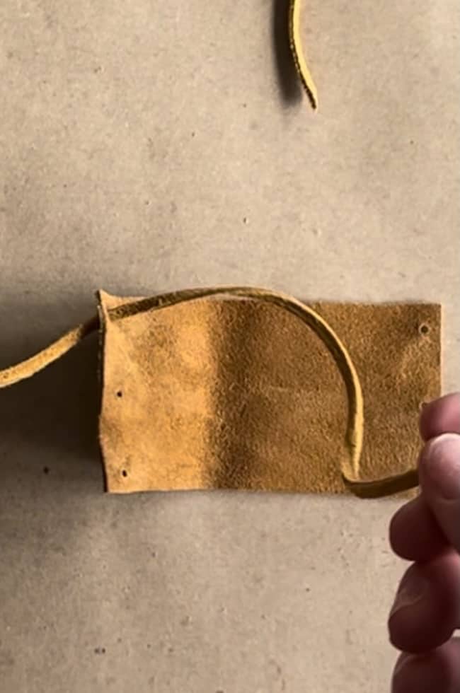 A close-up of a piece of brown suede leather with a hand holding a thin strip of the same material. The leather has a rough texture and small perforations on one side. It is set against a plain beige background.