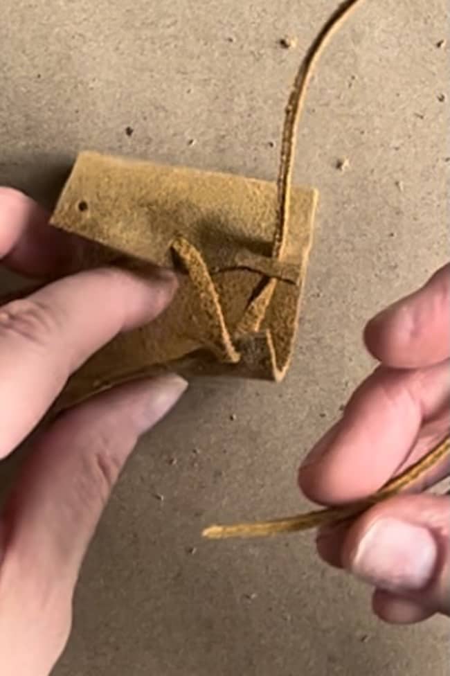 Two hands are crafting a small brown leather pouch on a cardboard surface. One strip of leather is being threaded through the pouch, demonstrating a weaving or assembling process.