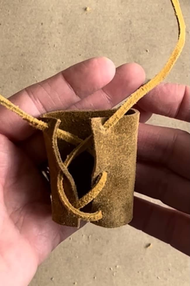 A hand holding a small, cylindrical, brown suede pouch with laces at the front. The background is a plain surface.