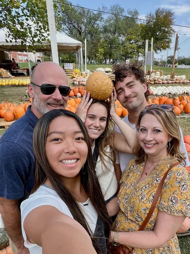 A group of five people standing in a pumpkin patch, smiling for a selfie. One person playfully holds a pumpkin on another's head. Various pumpkins are displayed in the background under a clear sky.