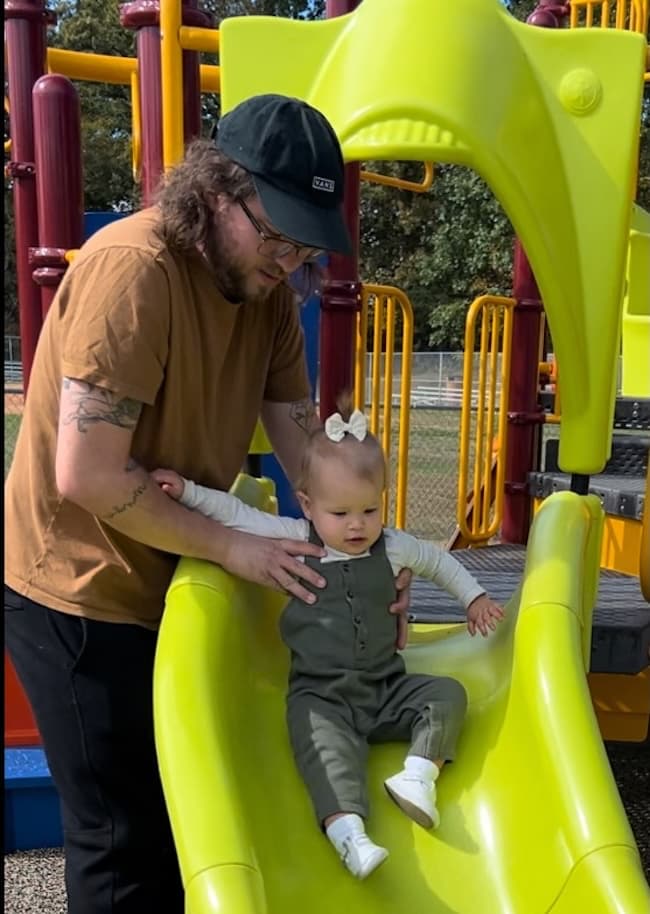 An adult helps a small child wearing a bow slide down a bright yellow slide at a playground. The playground equipment is colorful, and trees are visible in the background.