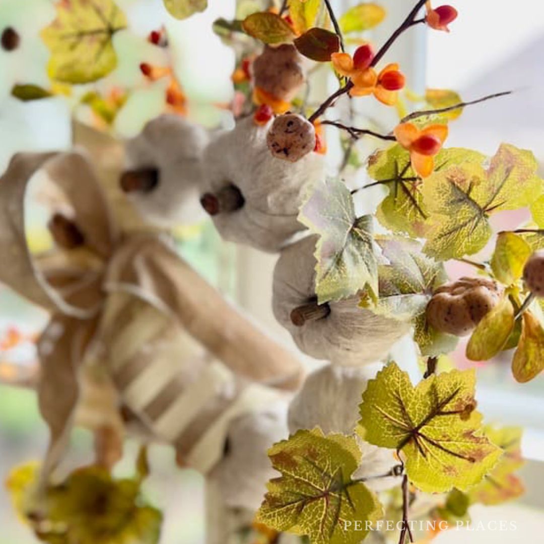 A decorative wreath featuring white cotton bolls, orange berries, and green-yellow leaves captures the essence of October 2024. A burlap ribbon is woven through, adding a rustic touch. The softly blurred background highlights the autumn-themed arrangement in perfect harmony for Seven on Saturday.