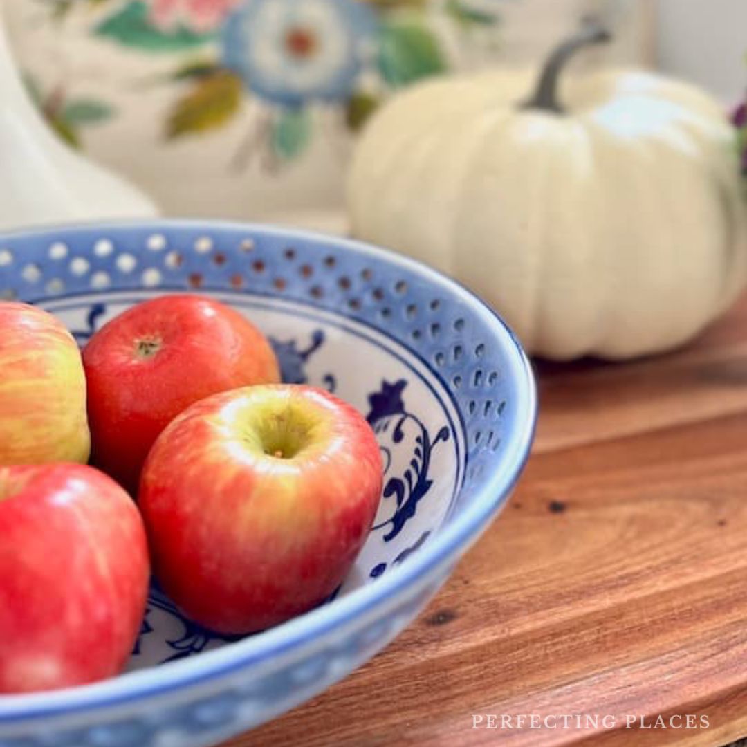 A blue and white patterned bowl filled with red and yellow apples sits on a wooden surface. In the background, a white pumpkin complements a floral cushion. Perfectly capturing the essence of October 5, 2025, this scene is reminiscent of a cozy Seven on Saturday moment.