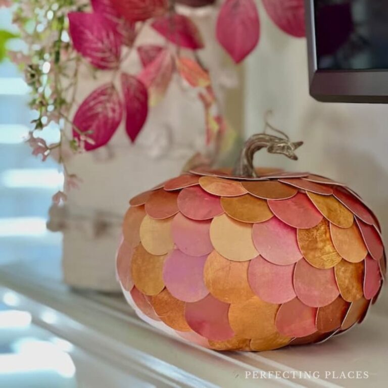 A decorative pumpkin with scales in shades of orange, pink, and gold sits on a white surface, showcasing an easy DIY pumpkin decorating technique using metallic paper circles. Behind it, there's a white vase with pink leaves and the edge of a black frame is visible on the right.