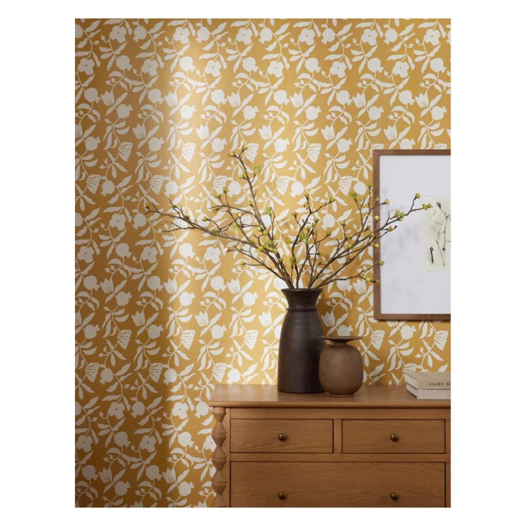 A wooden dresser with a black vase of branches and a small round pot sits against a yellow wall, showcasing the classic Sherwin Williams paint color palette with white floral patterns. A framed picture hangs on the wall, partly visible, creating a cozy and artistic setting.