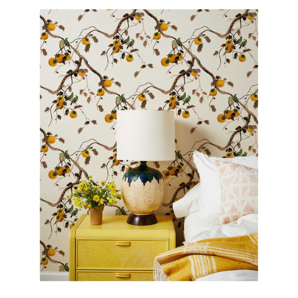 A cozy bedroom corner features a yellow bedside table and ceramic lamp, set against a wall adorned with floral and branch patterns. The classic Sherwin-Williams paint color palette complements the pastel bedding, while a small vase of flowers and a folded yellow-and-white blanket add charm.