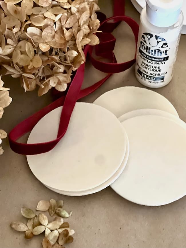 Wooden coasters, a bottle of white acrylic paint, dried hydrangea flowers, and a red ribbon are arranged on a beige surface.