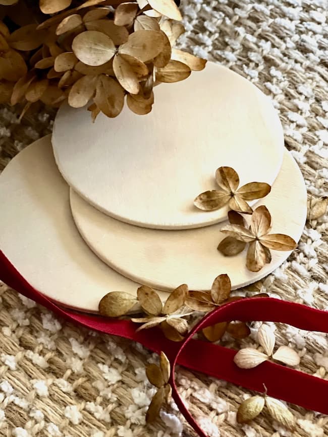 Close-up of three round wooden coasters stacked on a textured beige surface, surrounded by dried hydrangea flowers and petals. A red ribbon is partially visible in the foreground.