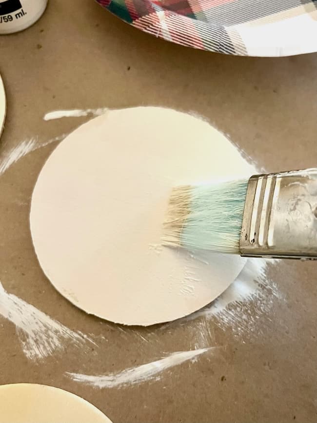 A flat, round white object is being painted with a light blue brush on a brown surface. A paper plate with a colorful pattern is partially visible in the background.