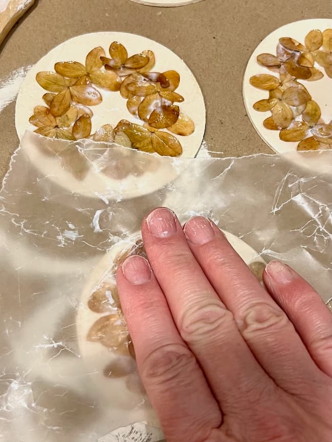 A hand pressing wax paper over a round piece of clay decorated with flat, petal-like objects in a circular pattern. The clay piece is on a flat surface with other similar pieces visible in the background.