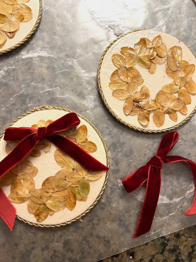 Three handcrafted ornaments feature dried flowers pressed onto circular bases. Two have red velvet bows; one is without a bow. The edges are decorated with a textured border, and they are arranged on a marbled surface.