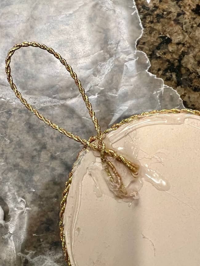 A candle ornament with a gold braided string loop is seen resting on a granite surface covered with crumpled foil. The candle has a shiny, smooth finish.