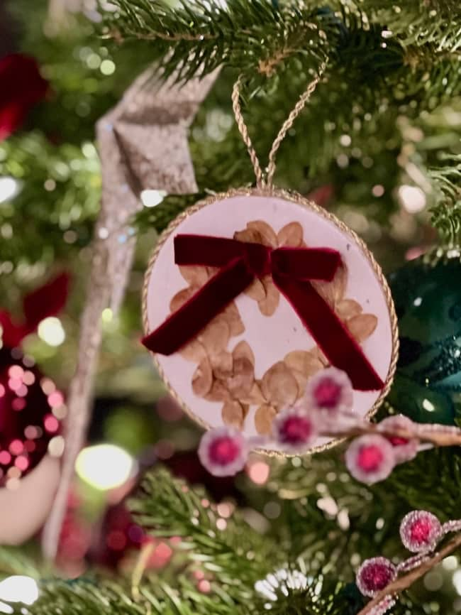 A close-up of a Christmas tree adorned with a handmade ornament featuring a circular frame, dried leaves, and a red velvet bow. The background includes blurred lights and various other colorful decorations.