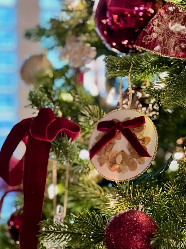 A close-up of a decorated Christmas tree featuring red velvet ribbons, a round ornament with dried flowers and a red bow, and shiny red baubles. The tree is adorned with twinkling lights, creating a festive and cozy atmosphere.