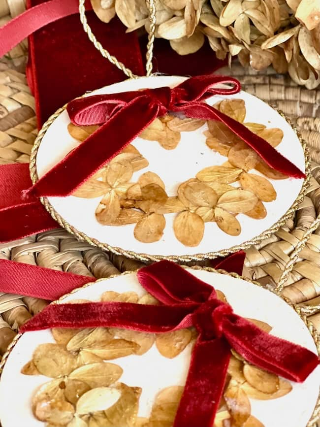 Two decorative round ornaments with dried flowers arranged in a circular pattern, adorned with red velvet bows. They rest on a woven surface with additional red ribbon visible in the background.