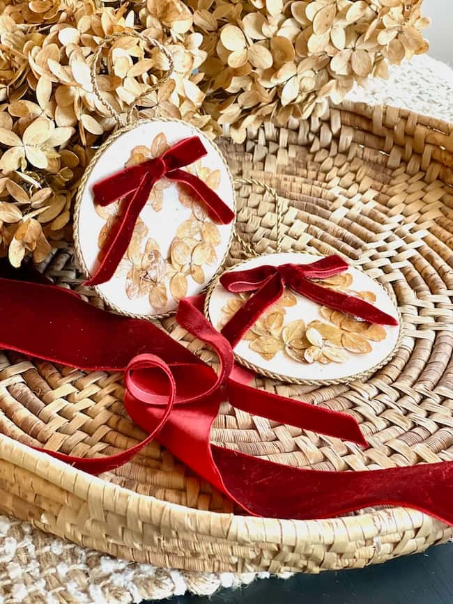 Two handmade ornaments with pressed flowers and red velvet bows rest on a woven basket. A red ribbon is arranged decoratively nearby. Dried hydrangea blooms are in the background, adding a rustic touch.